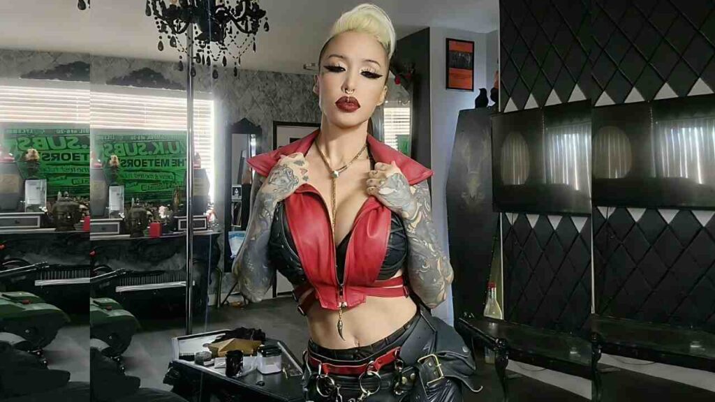 Masuimi Max with platinum updo and red lips in red cropped leather vest over black bra, posing by a pole in a studio.