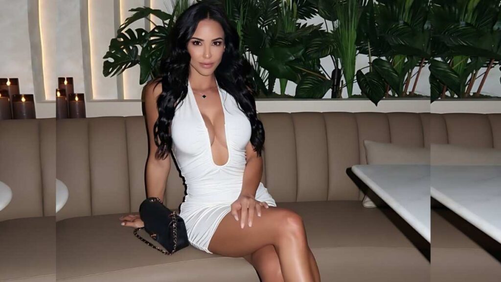 Melissa Riso in a white plunging dress sits on a beige booth, holding a black chain bag, candles and plants behind her.