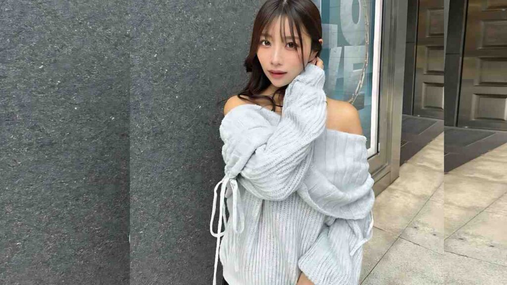 Mirei Kamiyama in a light-gray off-shoulder sweater leans against a stone wall, touching her face and looking at camera.
