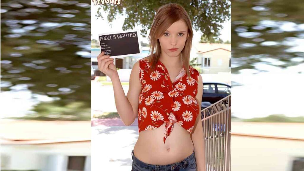 Missy Mae with light brown hair in a red floral tied crop top and jeans, holding a 'Models Wanted' sign outdoors.