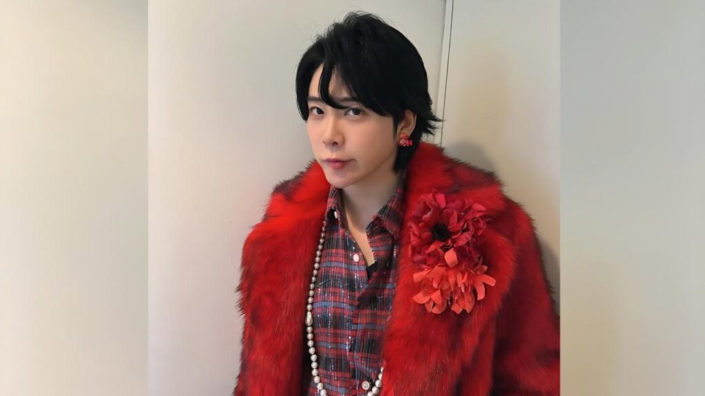 Motoki Ohmori with short black hair wearing a bright red faux fur coat, red plaid shirt, pearl necklace, and floral brooch.