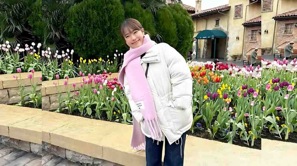 Nagisa Shibuya in a white puffer jacket, pink scarf and gloves poses beside a vibrant tulip garden along a stone path.