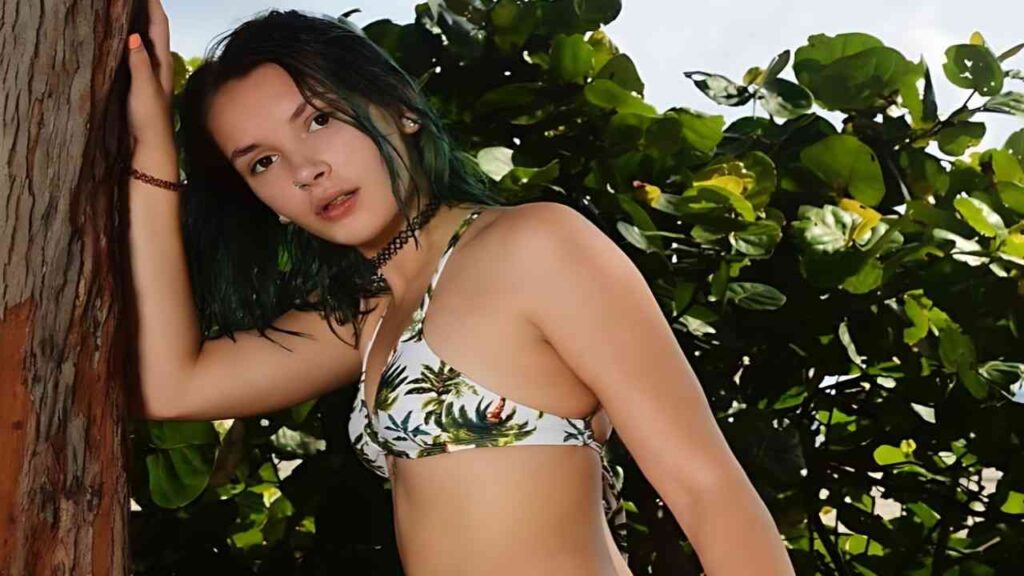 Natalie Porkman with teal-green hair in a palm-leaf bikini, leaning against a tree trunk beside lush green foliage.
