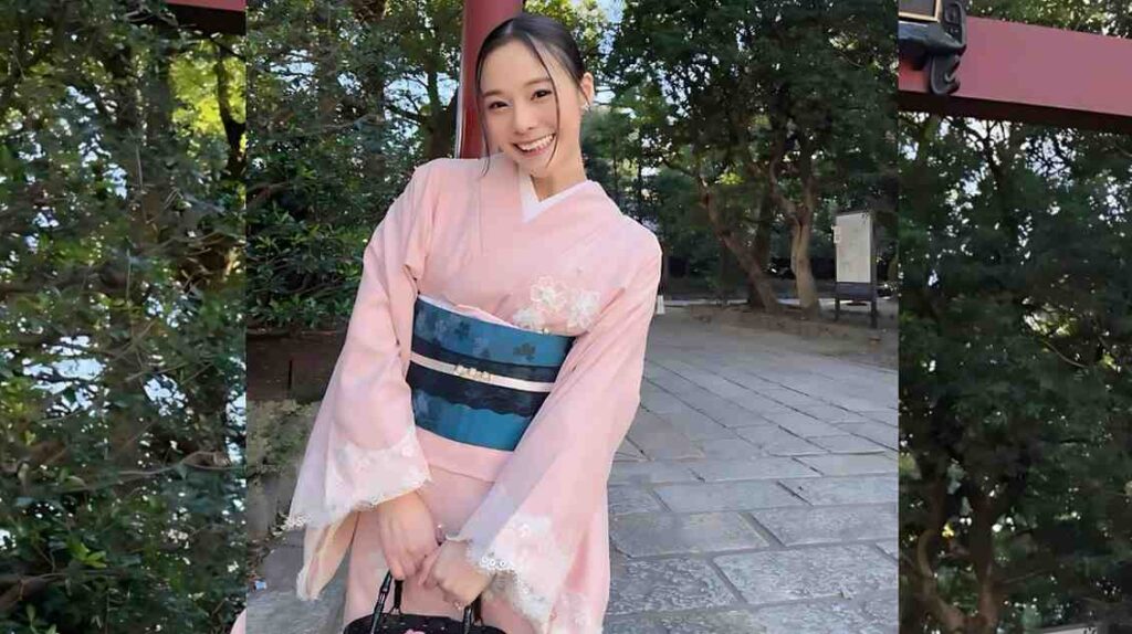 Nene Yoshitaka in a pale pink kimono with white embroidery and blue obi stands on a stone path among green trees.