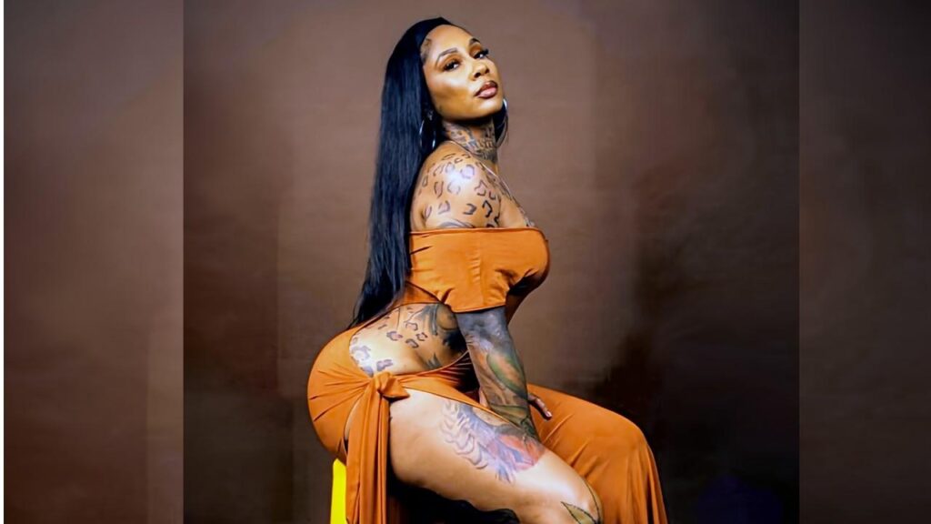 Nikki Sweets with long black hair in an orange off-shoulder dress, seated sideways, tattoos on arms and hip, brown backdrop.