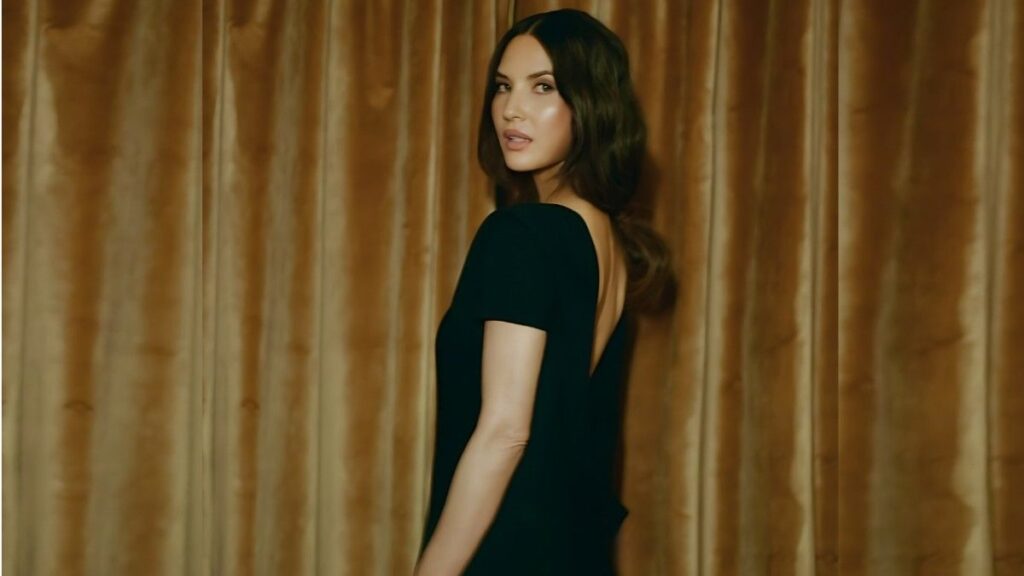Olivia Munn with dark hair in a black backless dress stands sideways before gold velvet curtains, gazing over her shoulder.