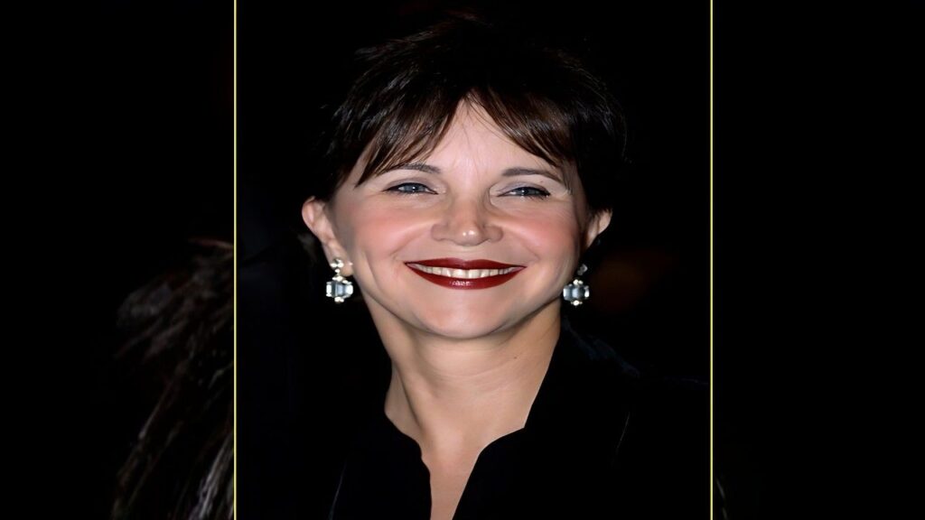 Priscilla Barnes with short dark hair, bold red lipstick, and diamond drop earrings, wearing black against a dark backdrop.