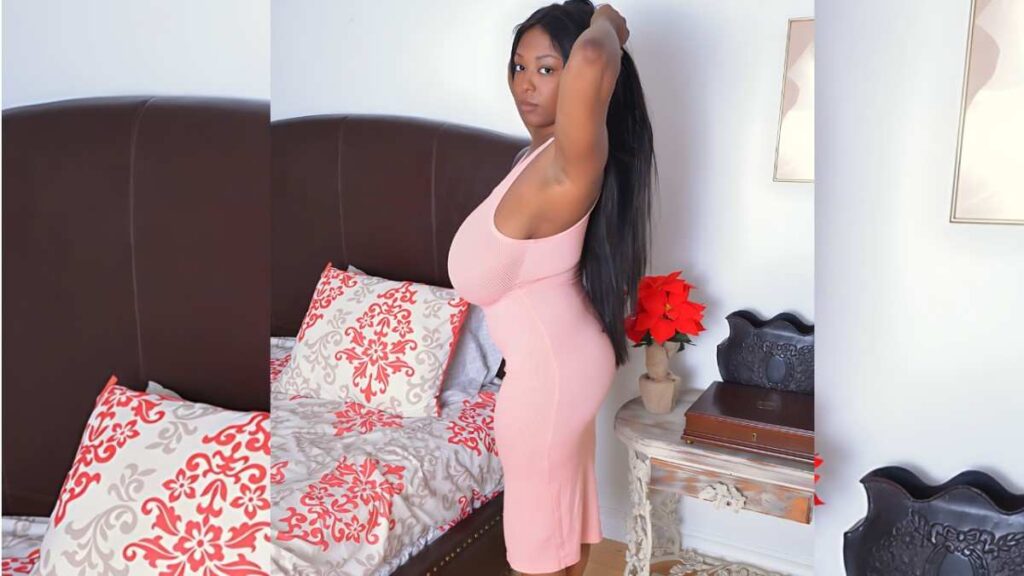 Rachel Raxxx in a pink bodycon dress stands beside a bed, adjusting her black hair; red floral pillows decorate the bed, and a side table holds a poinsettia.