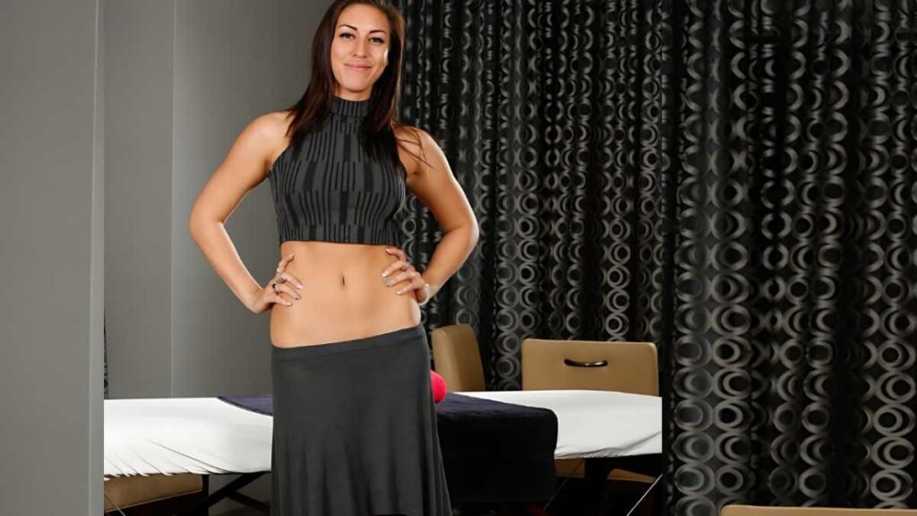 Rilynn Rae in a dark gray crop top and matching skirt stands with hands on hips in a room with a massage table.