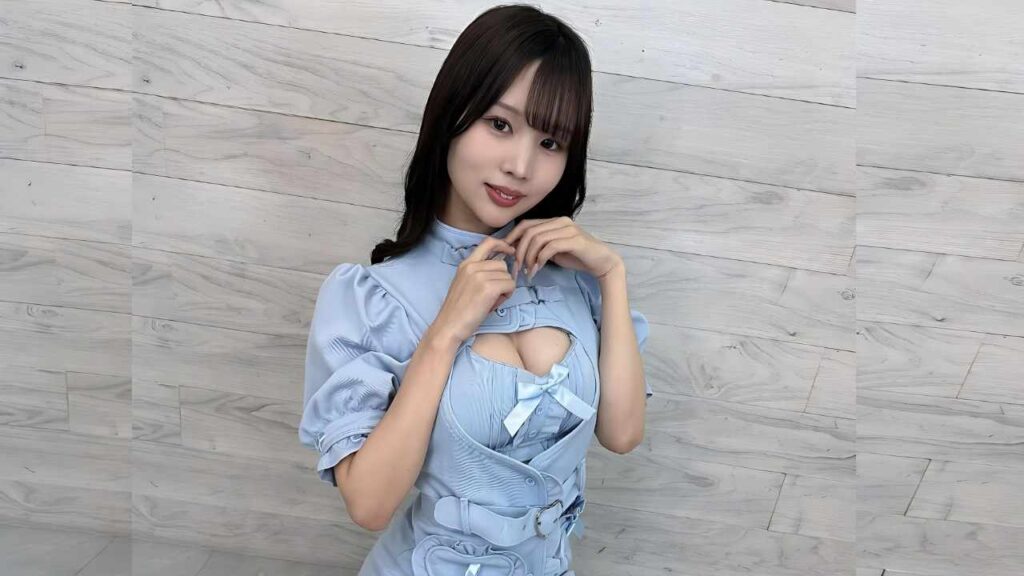 Rima Arai in blue puff-sleeve dress with a chest cutout and bow, posing against a light wood wall, hands at collar, smiling