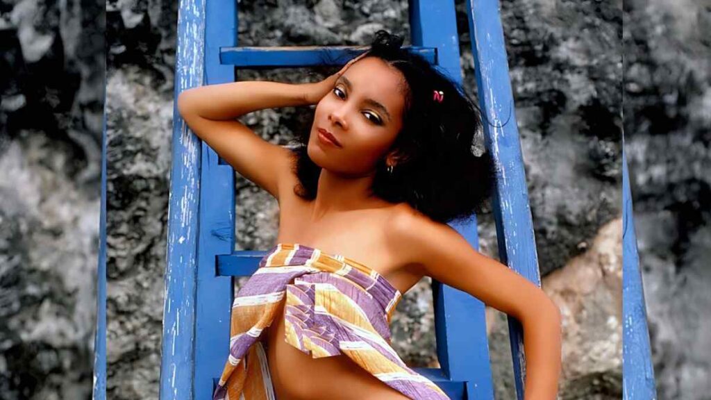 Rosanne Katon lounges on a blue ladder, arm resting behind her head, wrapped in a purple-orange wrap against a rocky backdrop.
