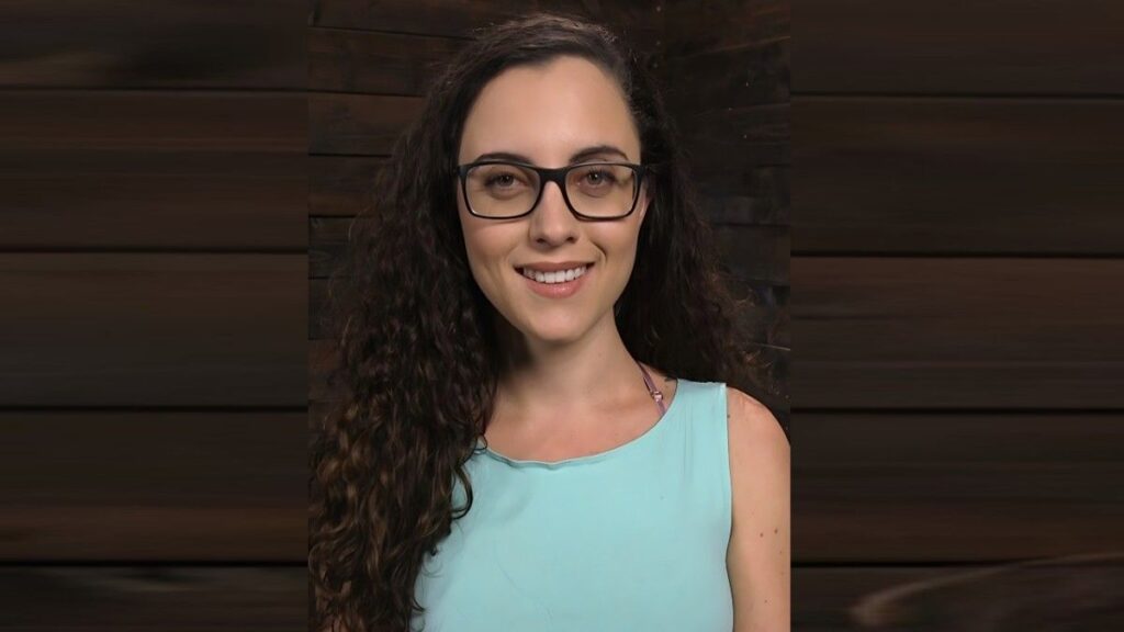 Roxanne Rae with long, curly hair and black-framed glasses, wearing a light blue sleeveless top, posed against wood panels.