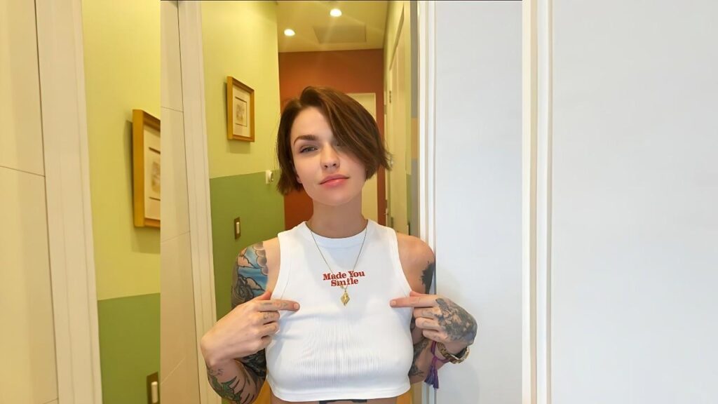 Ruby Rose with tattoos wears a white cropped tank and points to her shirt that reads Made You Smile, in a pastel hallway.