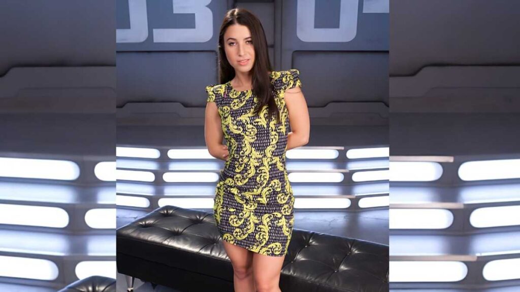 Serena Blair in a yellow and black patterned dress stands with hands behind her back in a futuristic studio with lights