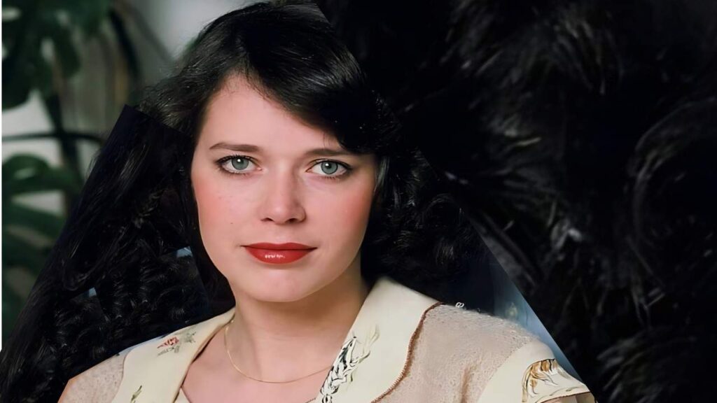 Sylvia Kristel in a beige floral blouse with embroidery, a thin gold necklace, and dark wavy hair, against a dark backdrop.