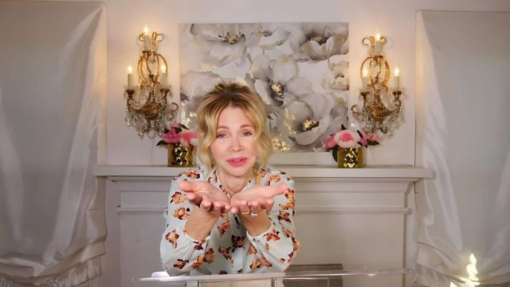 Teri Copley in a floral blouse stands at a clear lectern, cupping her hands toward the camera in a candlelit white set with flowers.