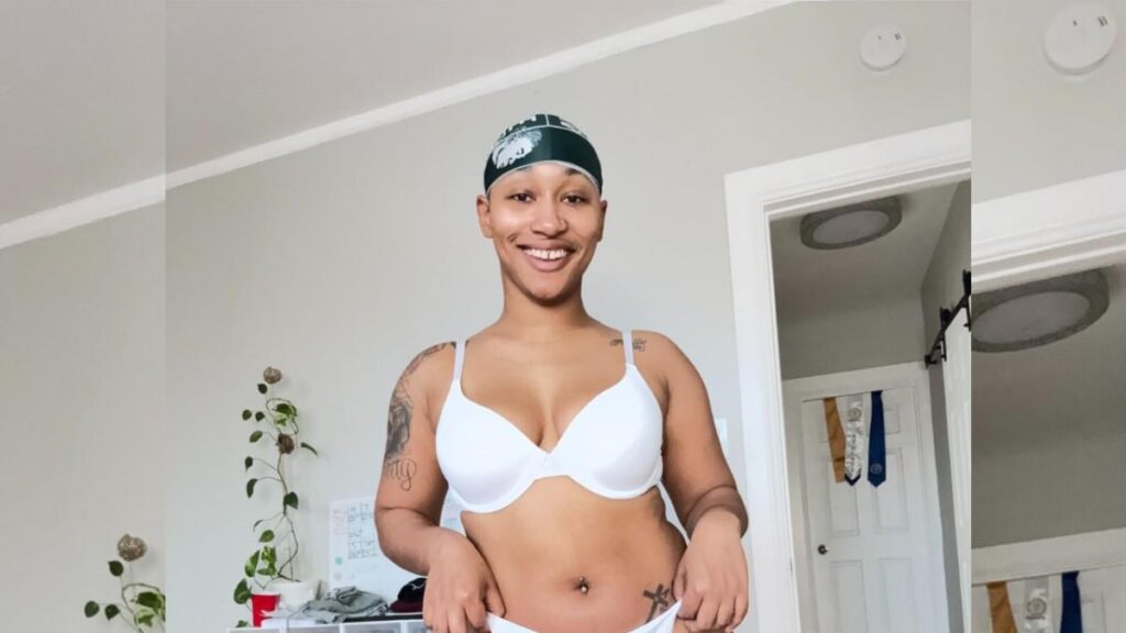 Thick Ass Daphne in a white bra and underwear with a green head wrap, tattoos visible, standing in a bright room near plants.