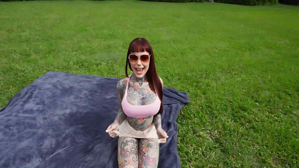 Tiger Lily with red hair in a pink tank top sits on a blue blanket on a green park lawn, smiling behind sunglasses.