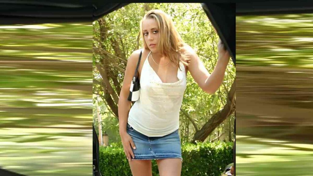 Tonya Harding in a white tank top and denim skirt stands by an open car door outdoors, purse over her shoulder, trees behind.