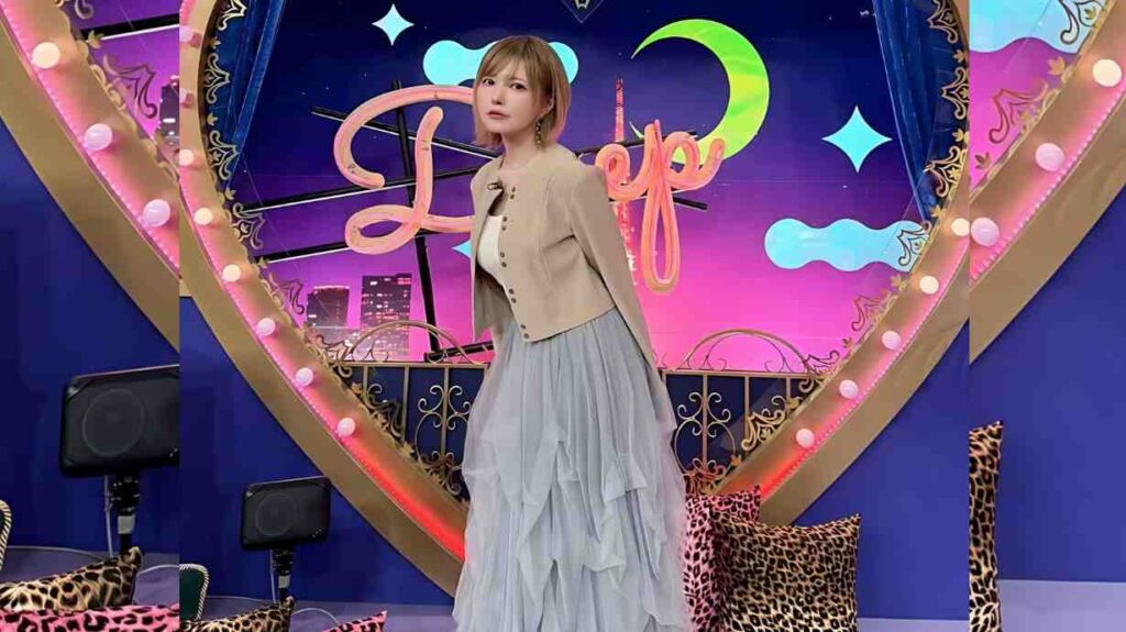 Tsubasa Masuwaka with short brown hair in beige cropped jacket and blue tulle skirt poses on a pink neon stage with gold arch