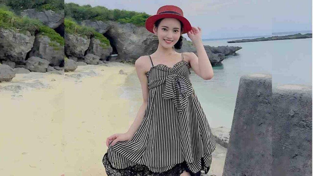 Umi Yatsugake in a black-and-white striped sundress and red hat poses on a sandy beach by rocks and calm turquoise water.