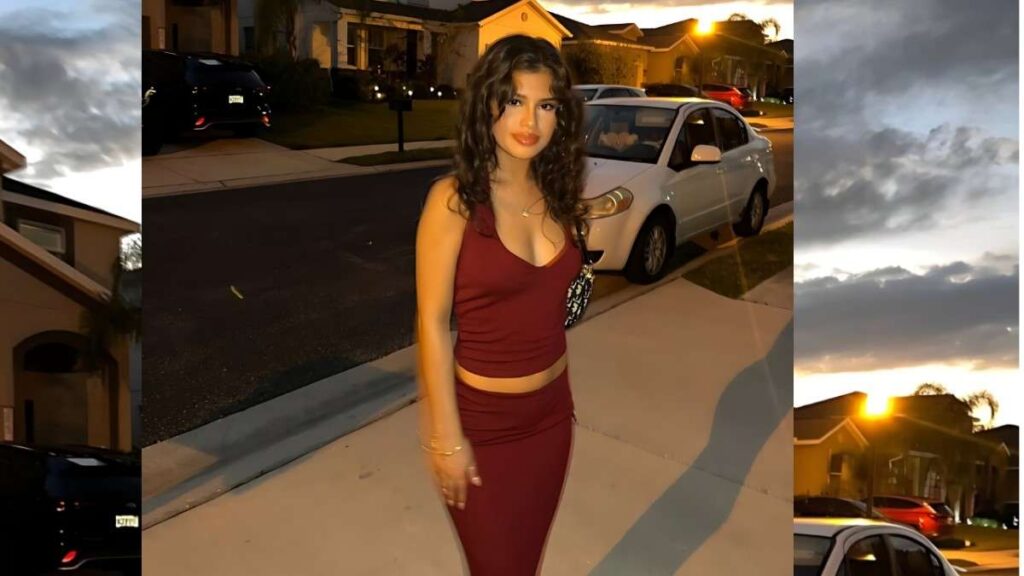 Victoria Jaimes with long, wavy dark hair in burgundy crop top and skirt stands on a suburban curb at dusk, bag over her shoulder, white car parked behind.