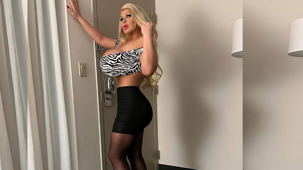 Victoria Vale in zebra print crop top and black high-waisted shorts poses by a hotel door, blonde wig and bold makeup.