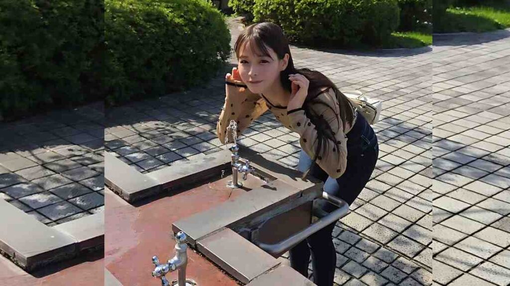 Yagi Nana with long dark hair leans toward a public water fountain on a tiled plaza, smiling in a beige cardigan and jeans.