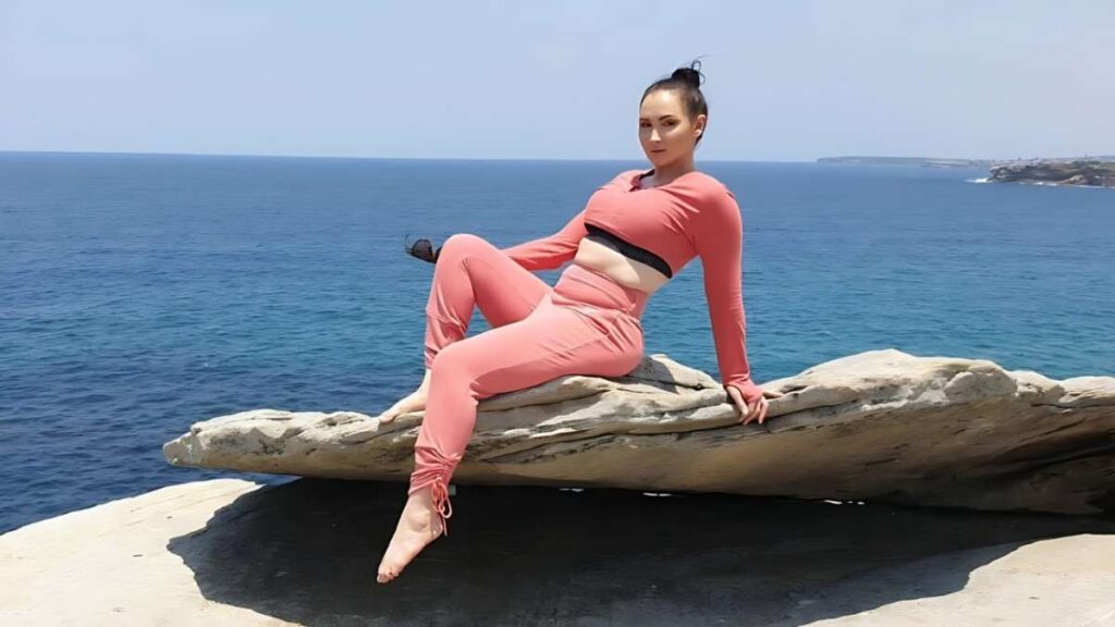 Yasmin Scott in coral-pink activewear sits on a flat coastal rock by the blue sea, legs relaxed and hair in a bun with distant coastline.