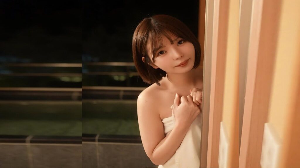 Yūka Ichii with a short brown bob, wrapped in a white towel, smiles softly by a doorway with beige curtains, warm lighting.