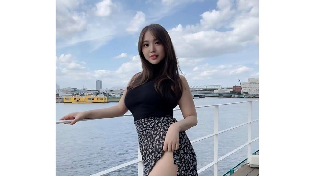 Yumi Mizusaki in a black sleeveless top and floral high-slit skirt leans on a railing by the water, with blue sky behind.