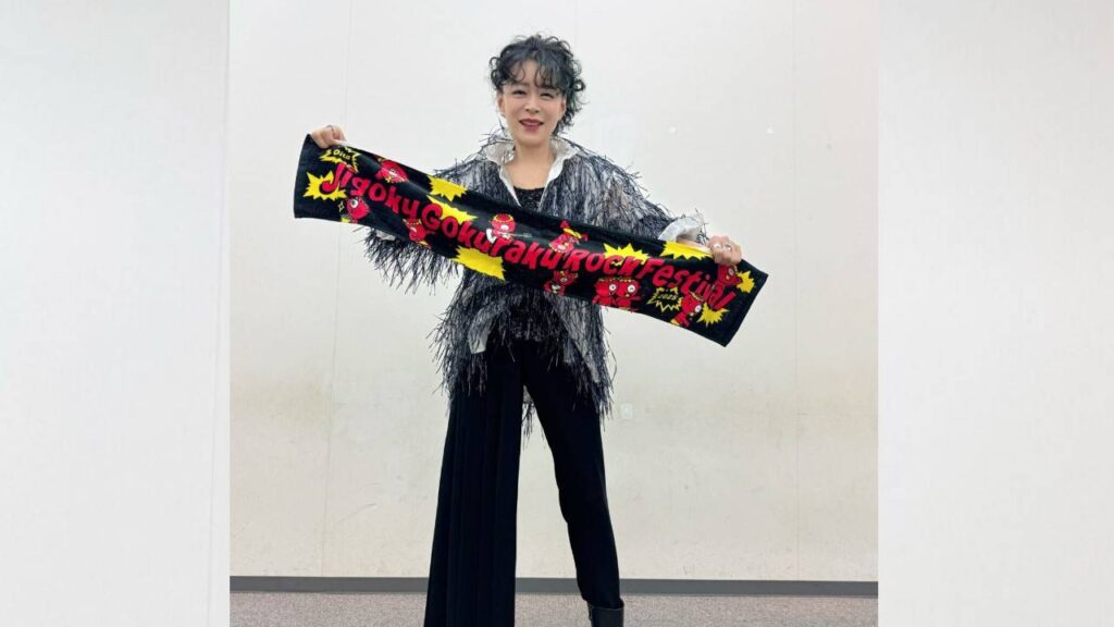 Akina Nakamori with short curly hair wears a metallic fringe jacket and black wide-leg pants, holding a colorful festival scarf.