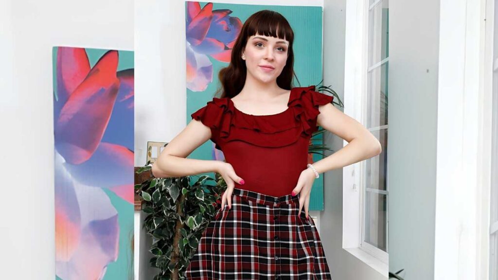 Aliya Brynn a dark bob and bangs wears a red ruffled top and red-black plaid skirt, posing in a room with florals.