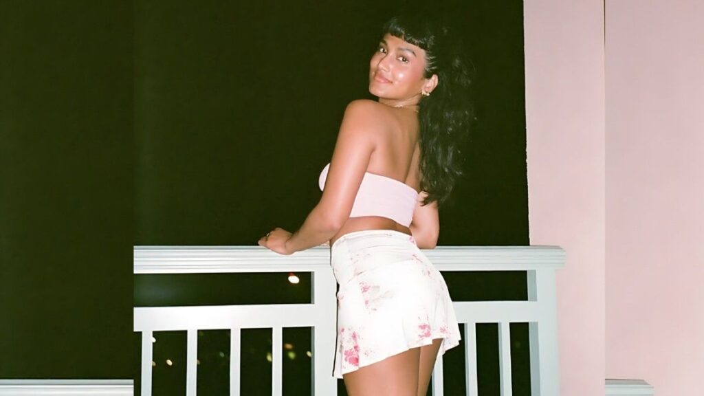 Angel Constance on a balcony at night, leaning on a white railing in a pink strapless top and white floral skirt, smiling.