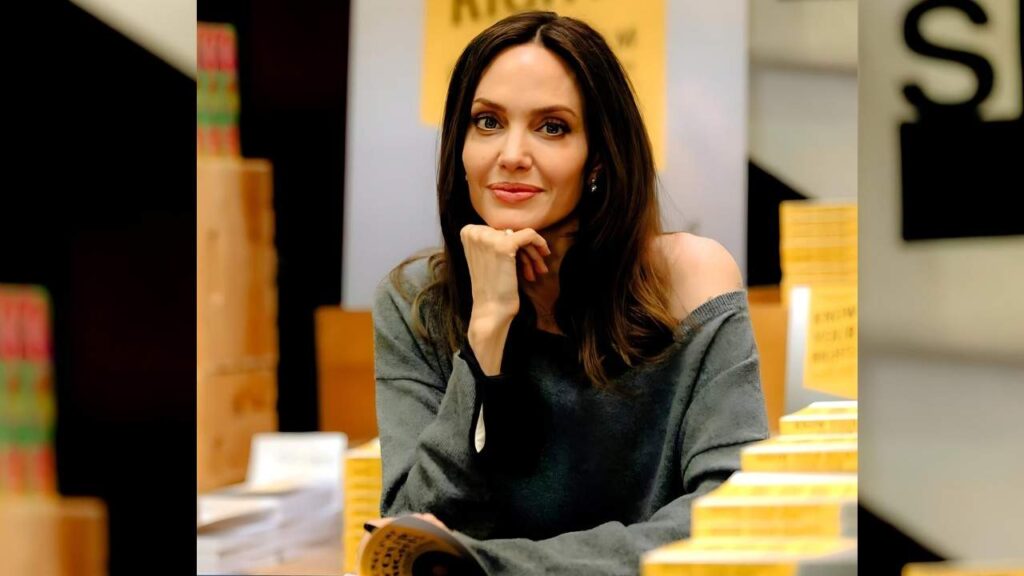 Angelina Jolie with long dark hair in a gray off-shoulder sweater rests her chin on her hand, smiling amid stacks of yellow books in a bright bookstore setup.