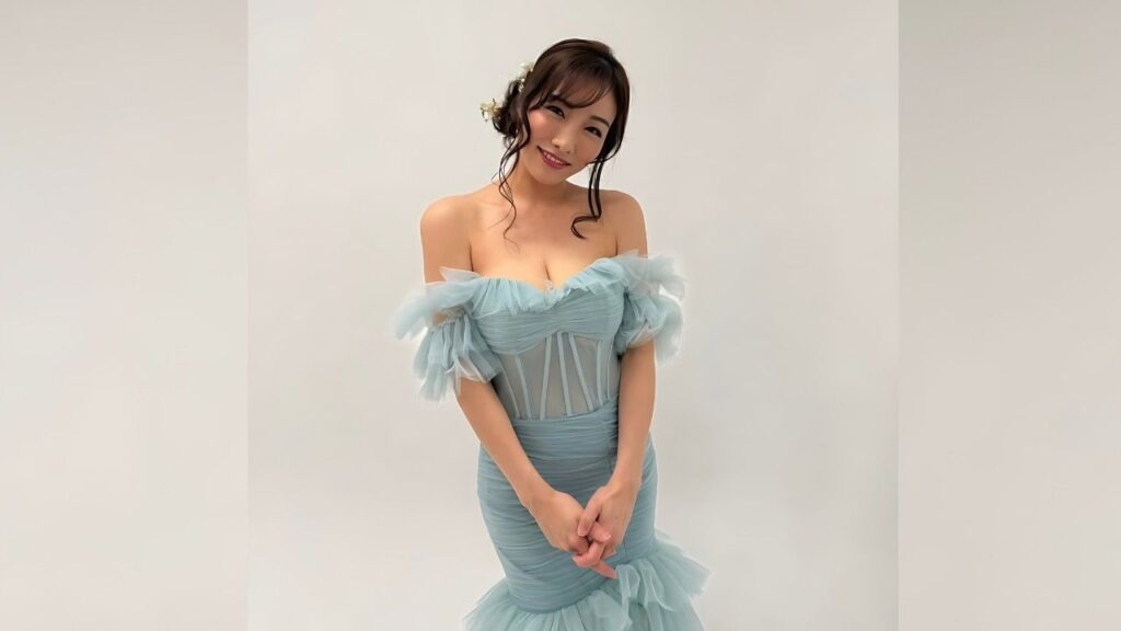 Anna Konno in an off-shoulder pastel blue tulle gown with ruffled sleeves, smiling and clasping hands in front.