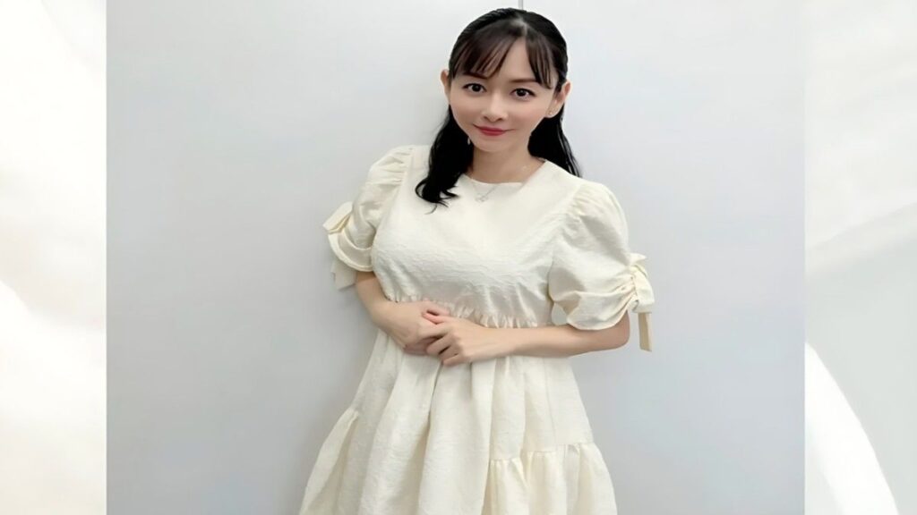 Anri Sugihara with dark hair in a cream puff-sleeve dress stands against a pale backdrop, hands gently clasped and a soft smile.