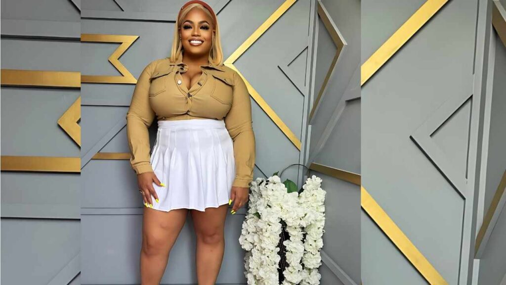 Ashley Renee in a tan button-down top and white pleated mini skirt stands against a gray geometric wall with gold accents.