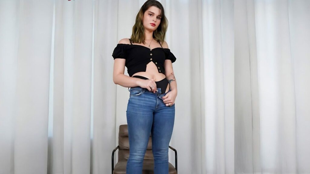Asia Belle with greenish hair stands before white curtains, wearing a black off-shoulder crop top and blue jeans.