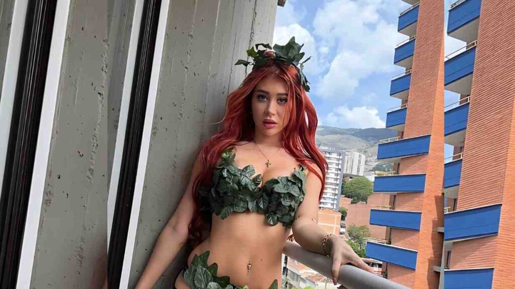 Atlanta Moreno in a leafy green bikini and crown, leaning on a balcony railing, with blue buildings and distant hills.