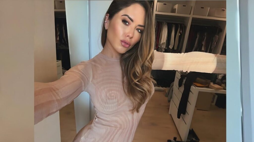 Brittney Palmer with long, wavy hair in a pale pink sheer top selfies in a bright walk-in closet filled with clothes.