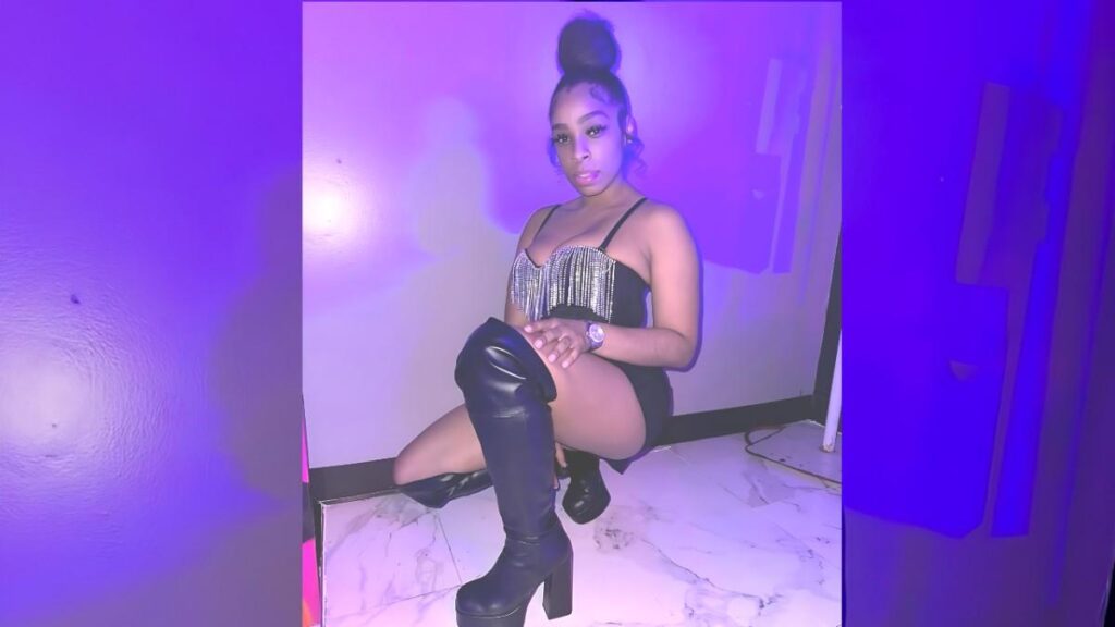 Brooklyn Lee in a neon purple-lit room, wearing a fringed black top, black thigh-high boots, a high bun, and a watch.