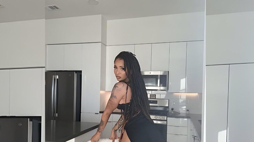 Cali Sweets with long braids and tattoos in a black dress leans on a white kitchen island amid cabinets and stainless appliances.