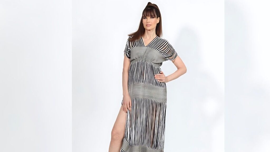Carol Alt in a black-and-white striped maxi dress with gray panels poses in a bright studio, hand on hip, side slit.