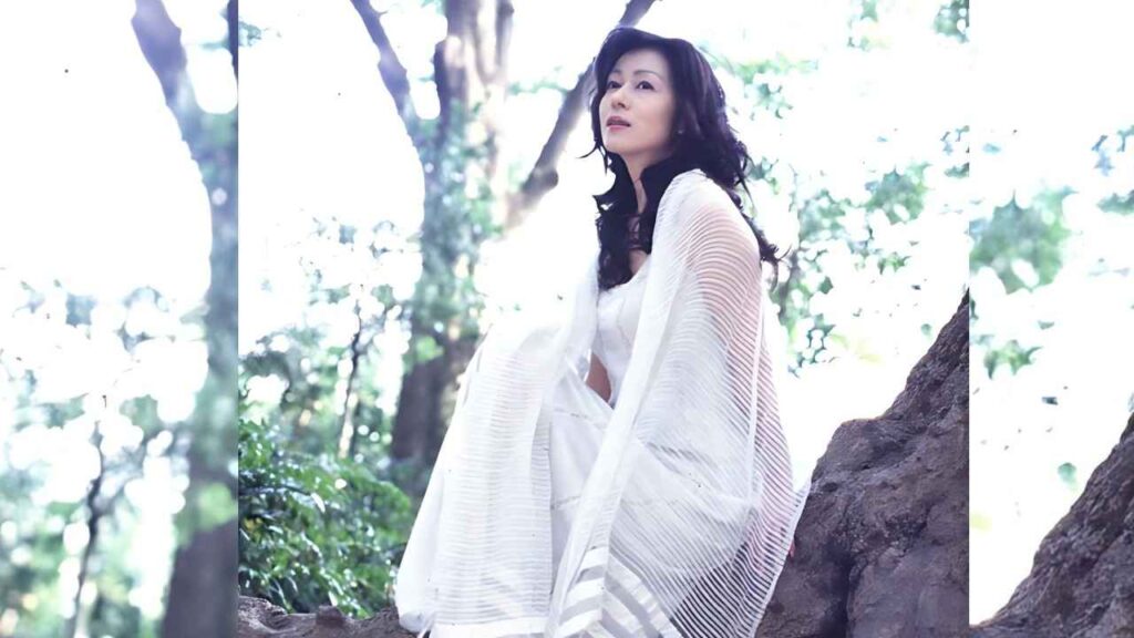 Chikako Aoyama in a flowing sheer white shawl sits on a rugged rock in a sunlit forest, her dark wavy hair framing a calm gaze.