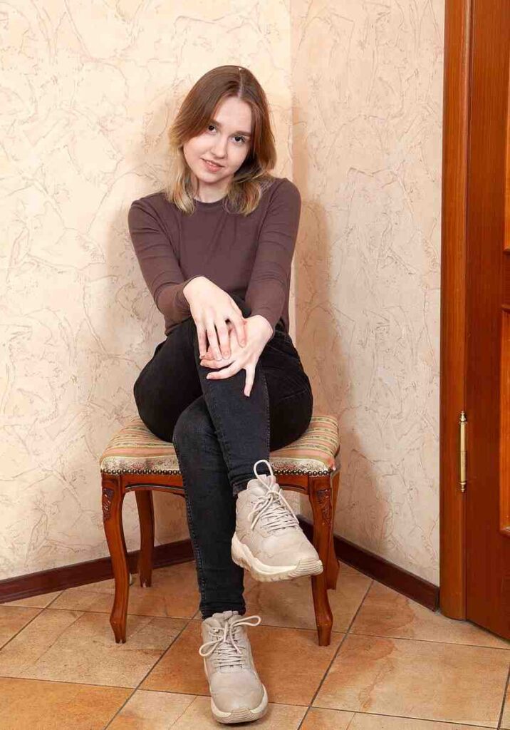 Doll Hole sits cross-legged on a carved wooden chair with striped upholstery in a peach room, wearing brown top and beige sneakers.