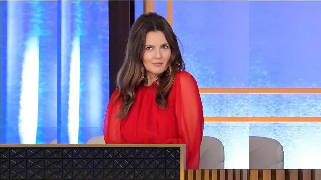 Drew Barrymore in a sheer red blouse sits at a desk on a bright TV studio set with blue panels and orange lines.