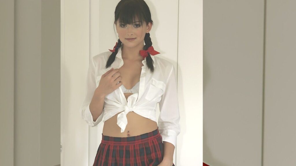 Ellie Idol with two braids and red ribbons wears a knotted white shirt exposing midriff over a red plaid skirt, in a doorway.