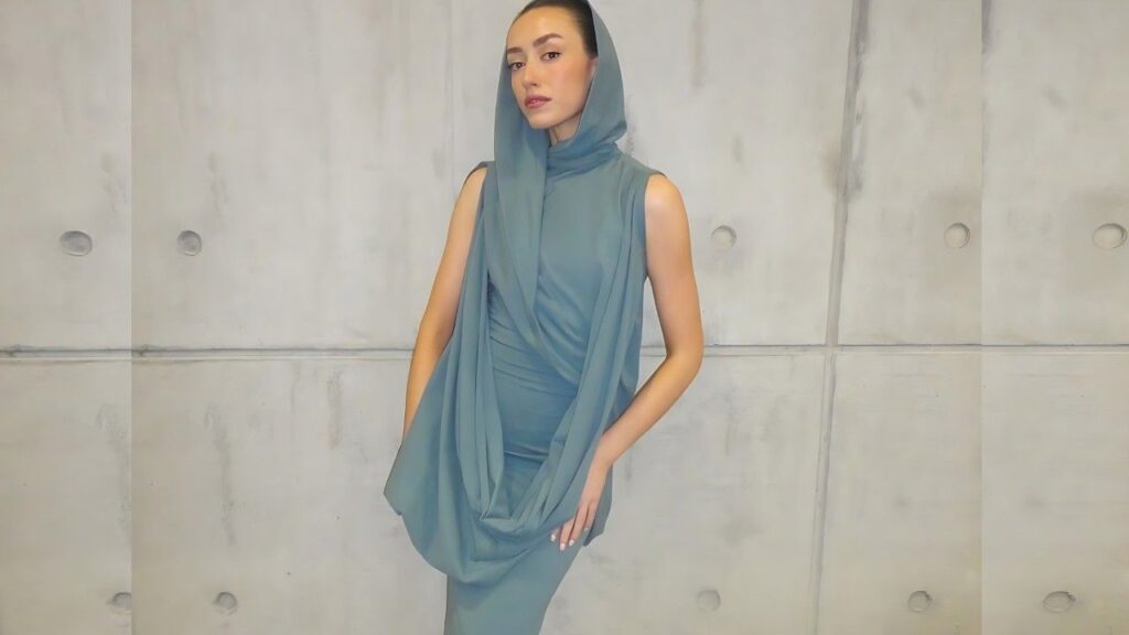 Ellie Roe in teal draped dress and hooded scarf poses against a pale concrete wall, hands on hips, calm expression.