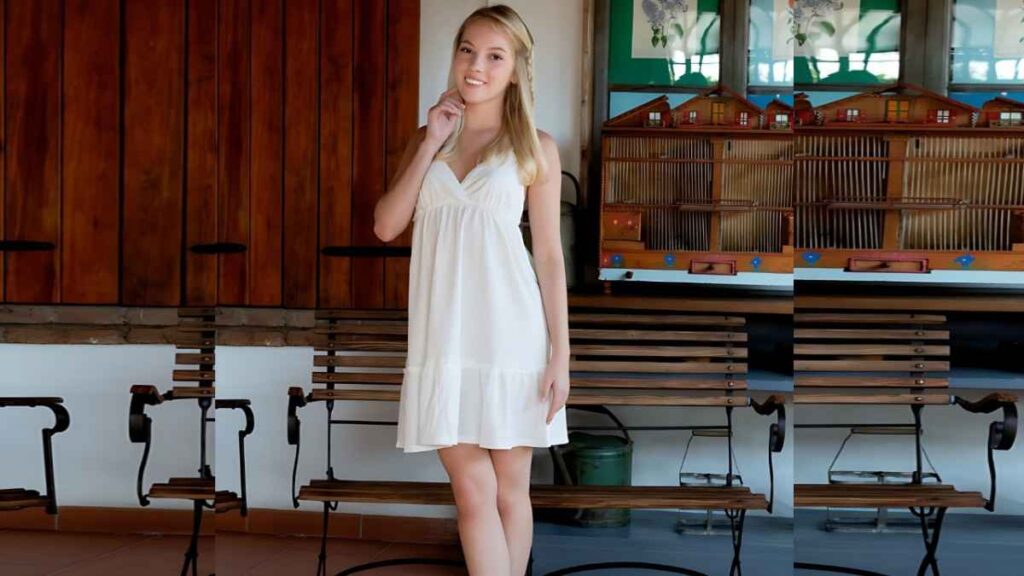 Emma White in a white sundress stands in a rustic room with wooden benches, smiling and touching her neck.